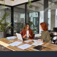 stock-photo-two-busy-business-women-of-young-and-middle-age-talking-in-creative-green-office-sitting-at-desk-2432308009